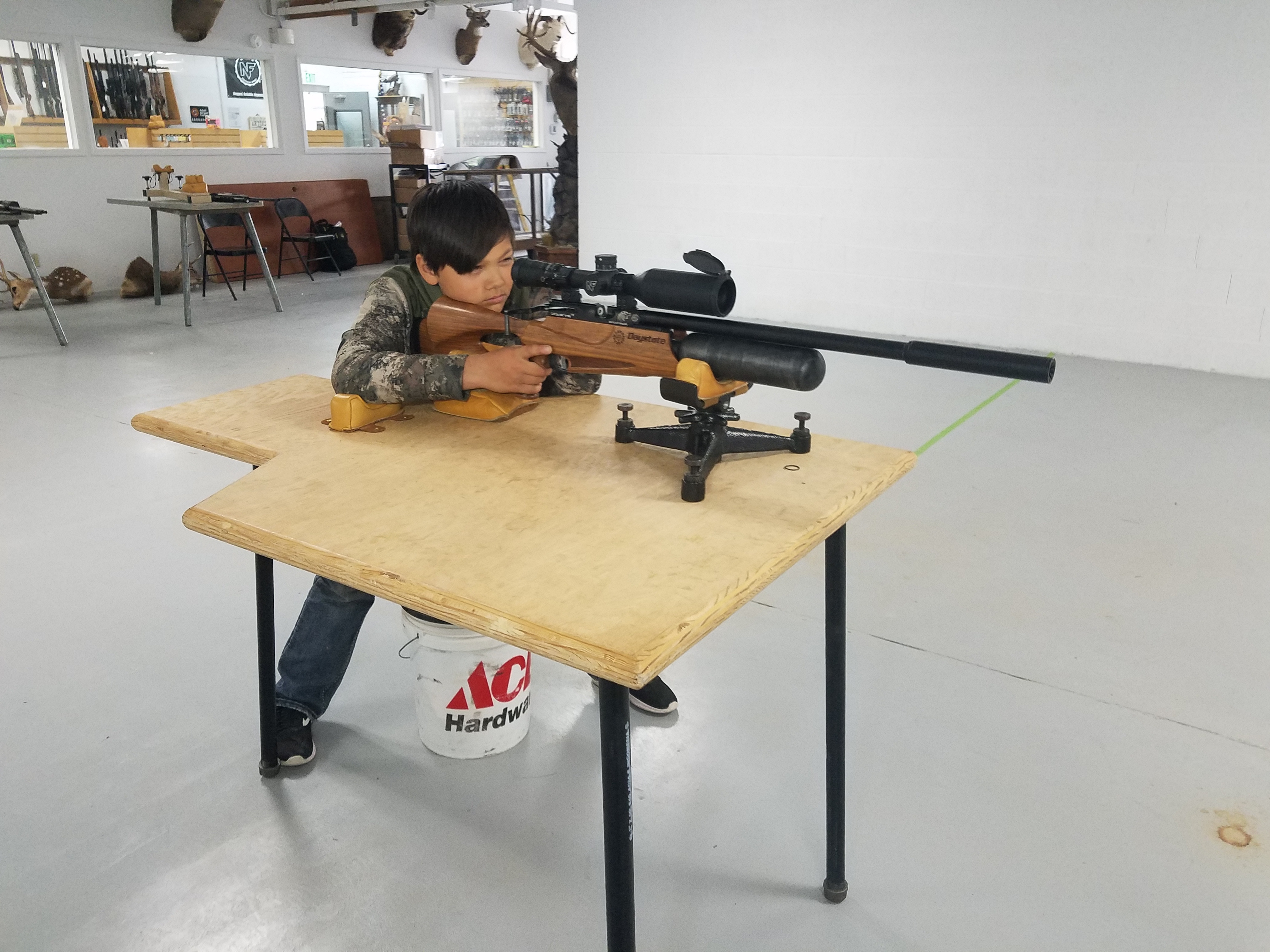 A young shooter is seated at table while he aims an air gun towards a target