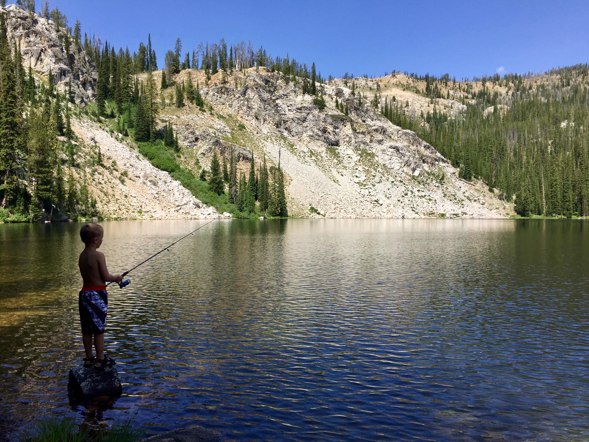 A boy wearing a swim suit fishes at an alpine lake.