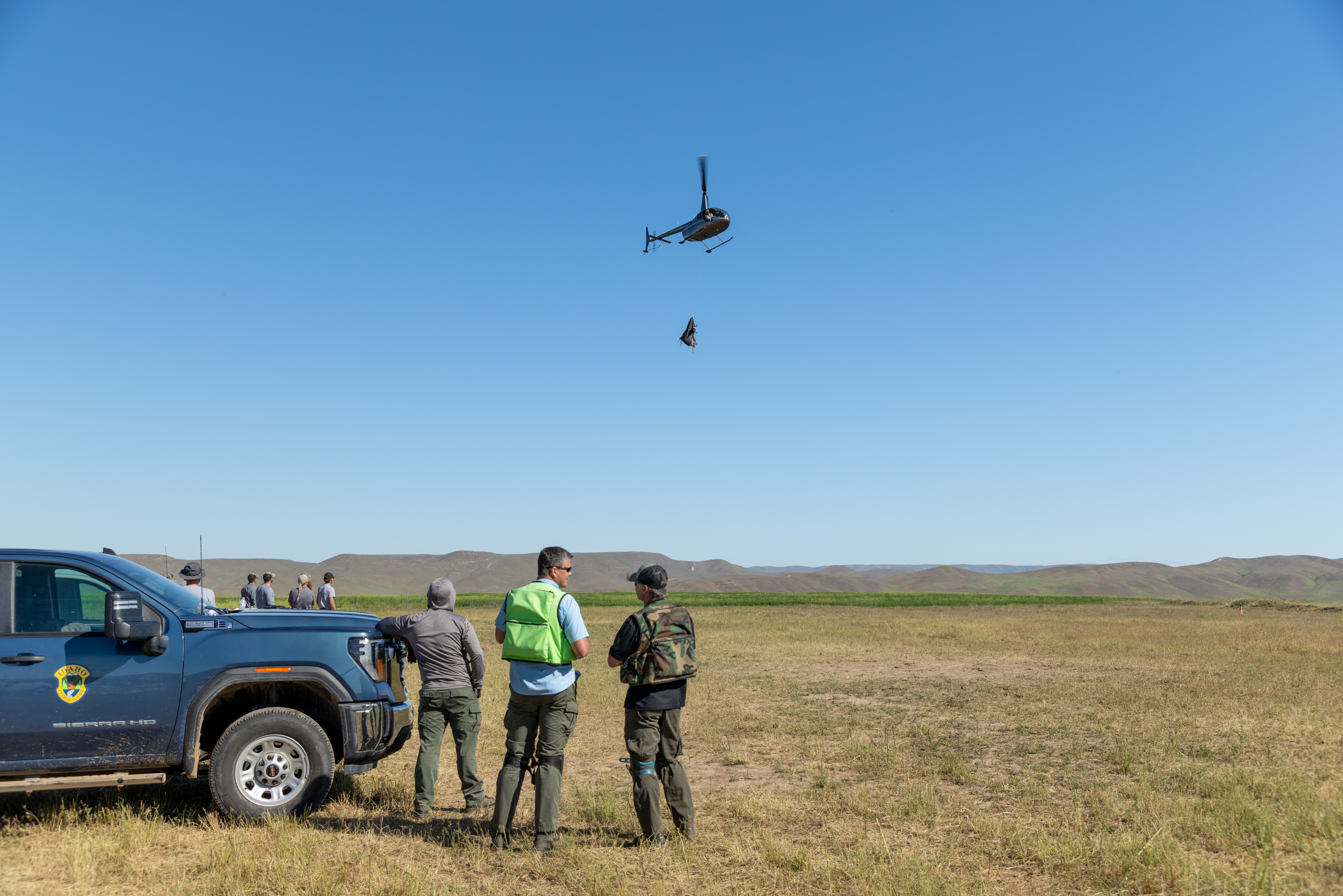 A helicopter, with an elk slung beneath it, travels toward IDFG staff standing by a truck on the ground.