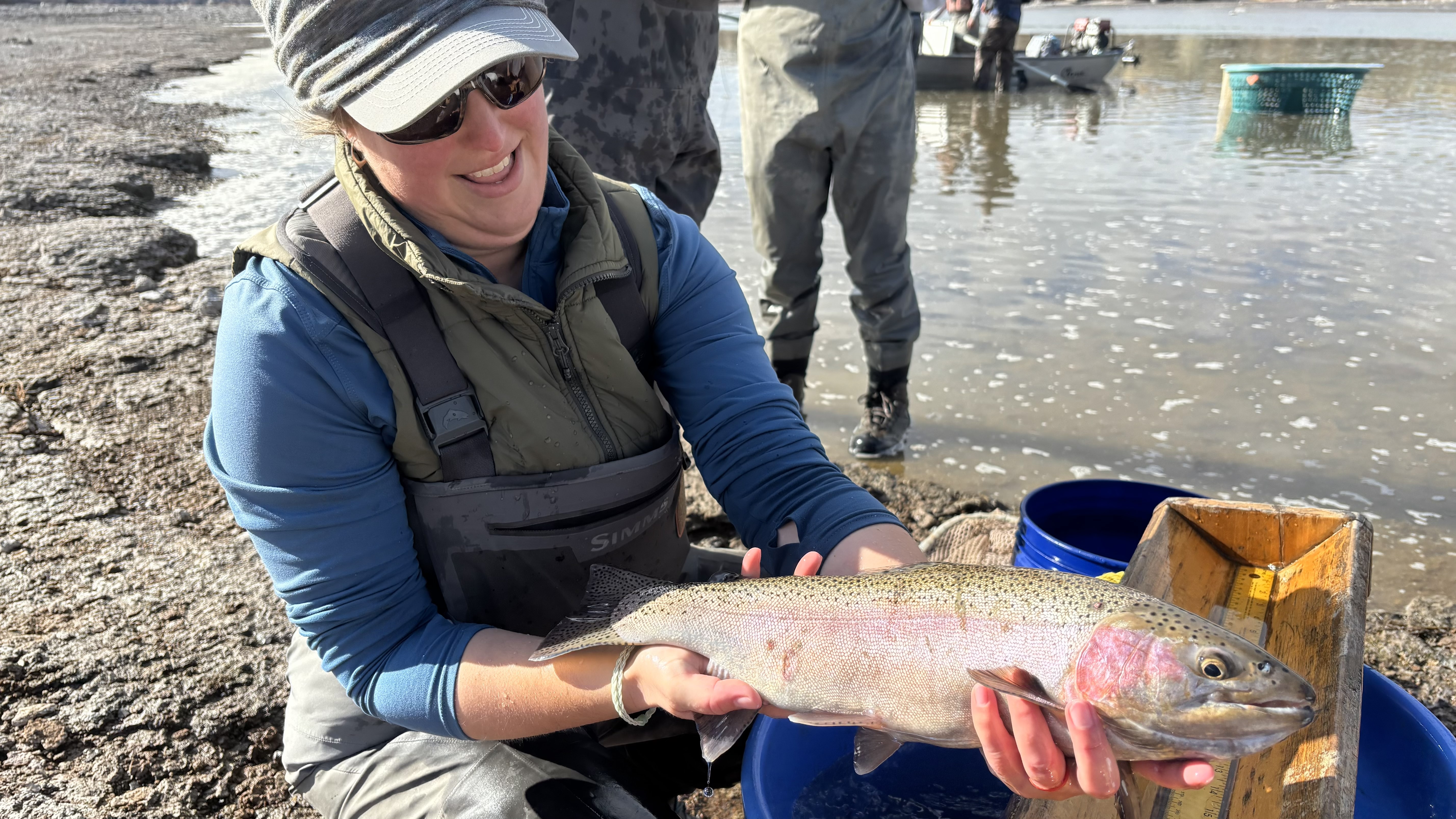 A woman in waders and cap holding a large rainbow trout with water and boat in the background
