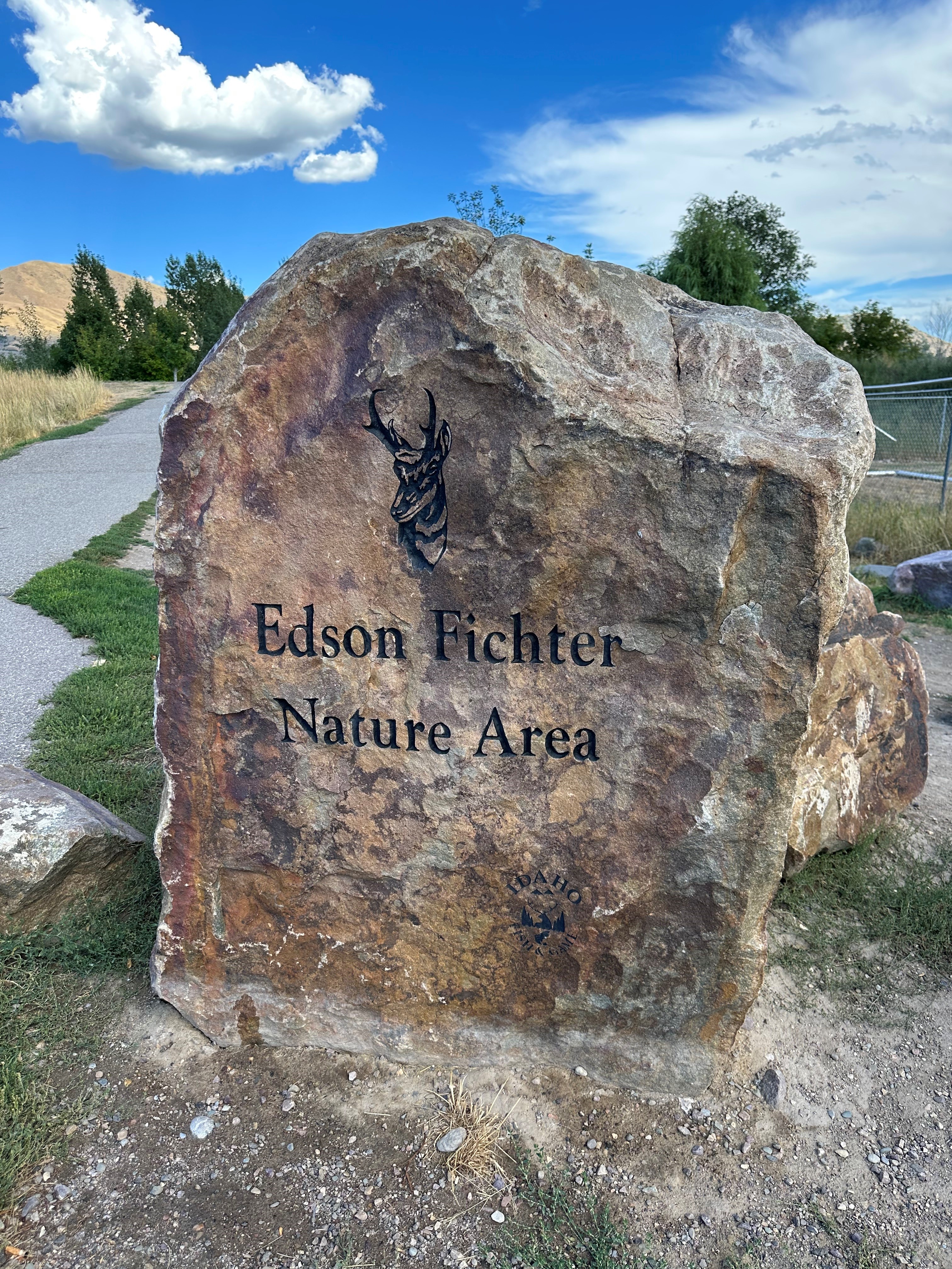 A large natural boulder engraved with the words Edson Fichter Nature Area is positioned at the entrance and next to a paved path at the Edson Fichter Nature Area.