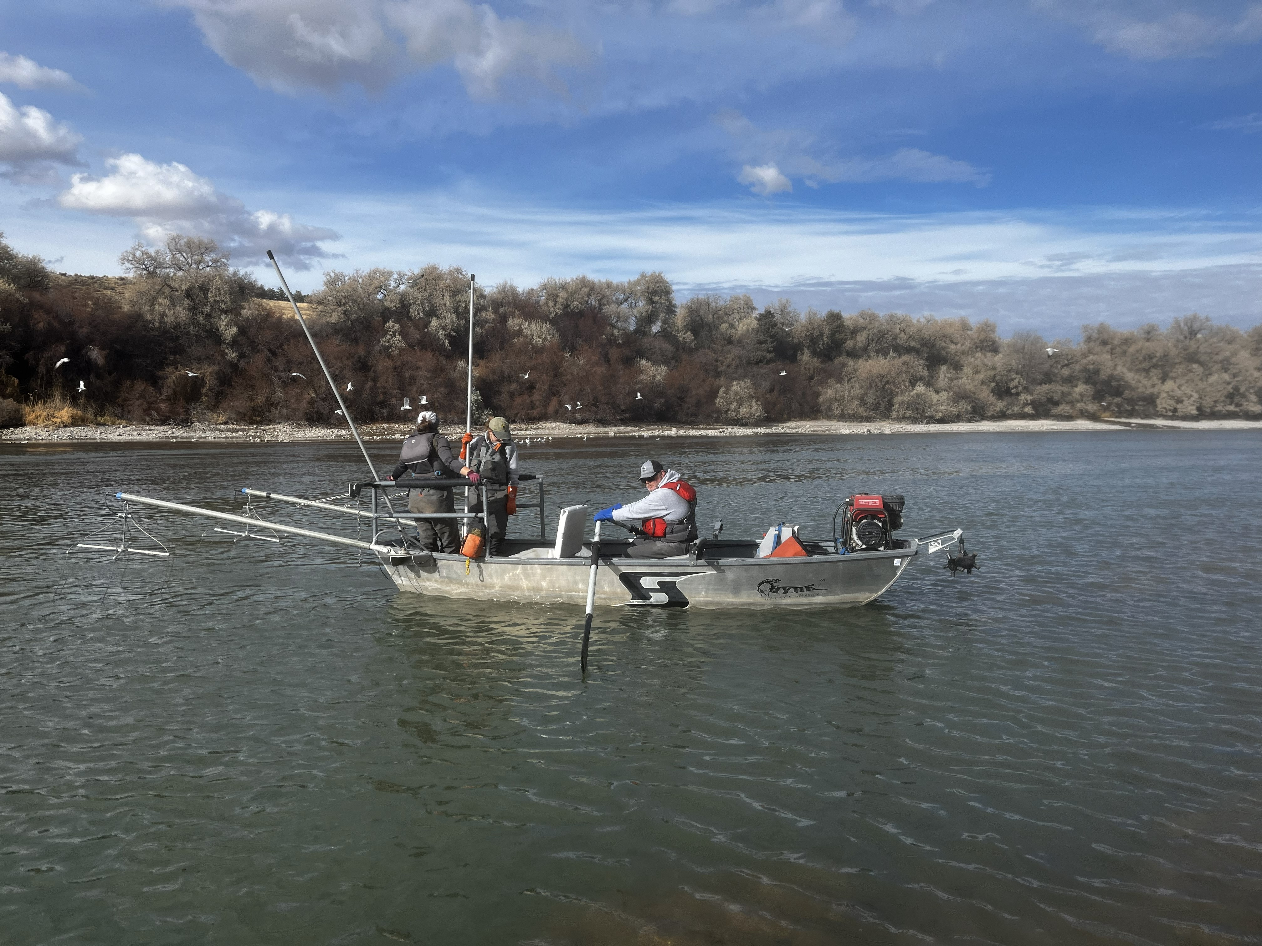 People in a drift boat on a slow-moving river with vegetation and a cloud-filled sky in background