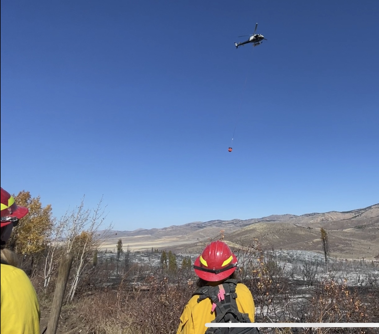 Two people in yellow shirts and red hardhats watch a helicopter drop water on a wildlfire