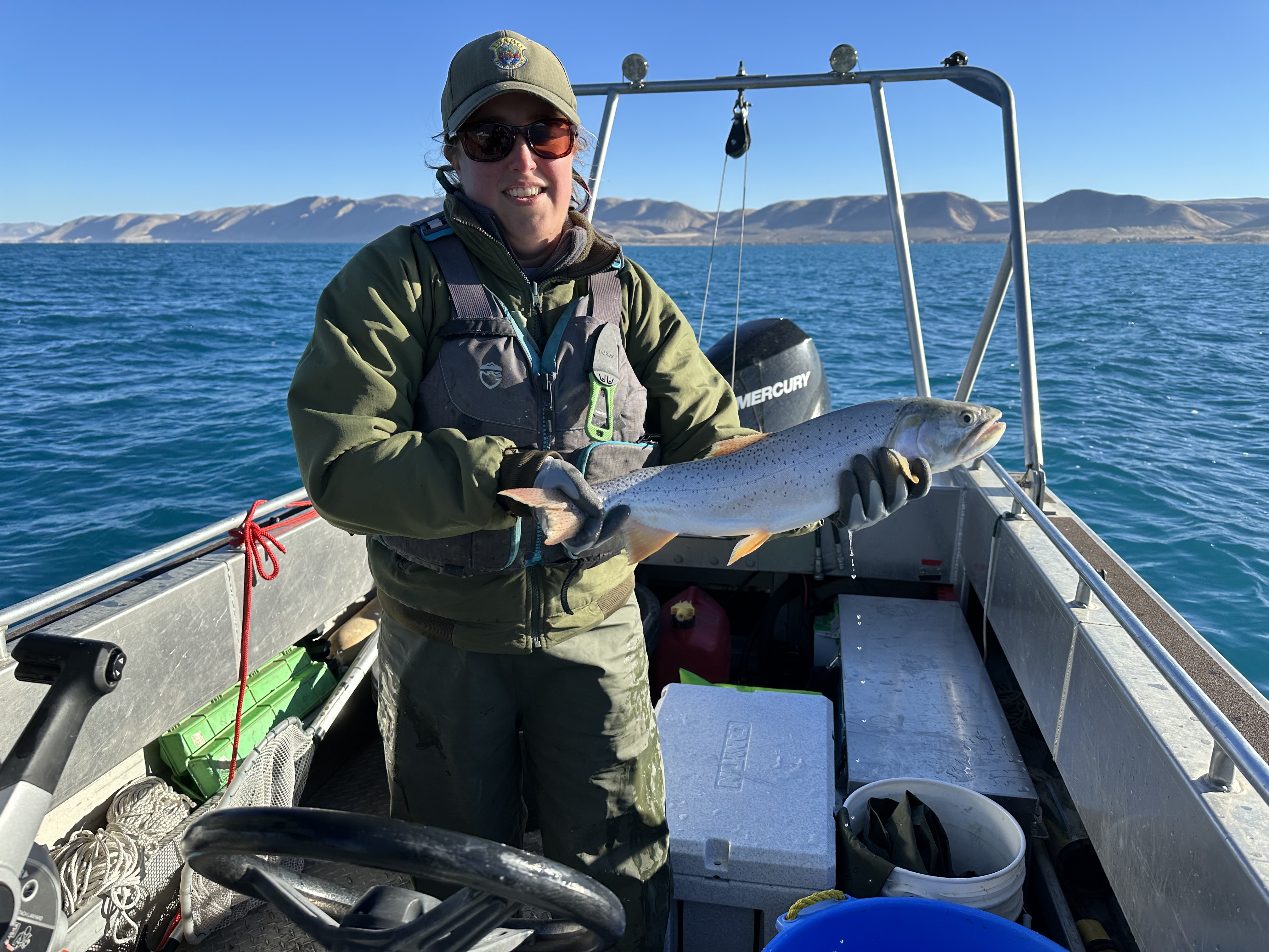 Female wearing a baseball cap and outdoor wear holds a fish while standing in a boat on a blue lake.  Distant mountains in the background.