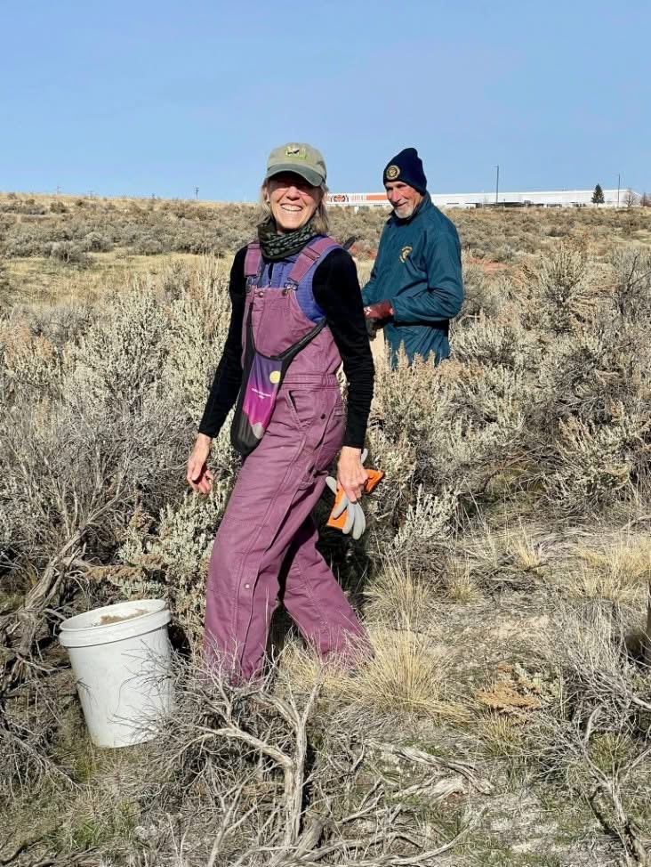 woman in foreground in pink coveralls standing amongst sage brush with man in blue jacket and stocking cap in background