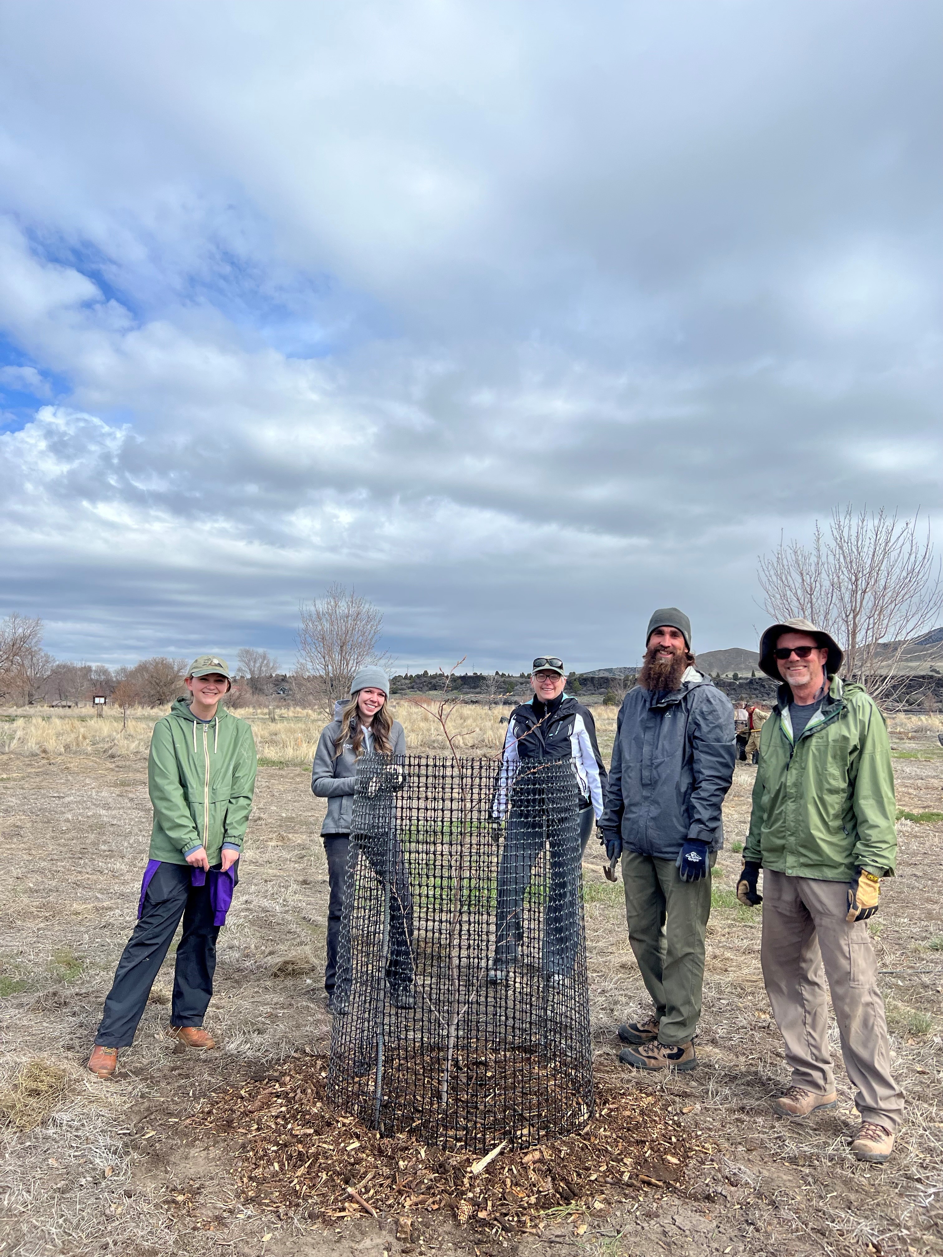 People standing behind a newly planted tree in an open area under a cloudy sky.