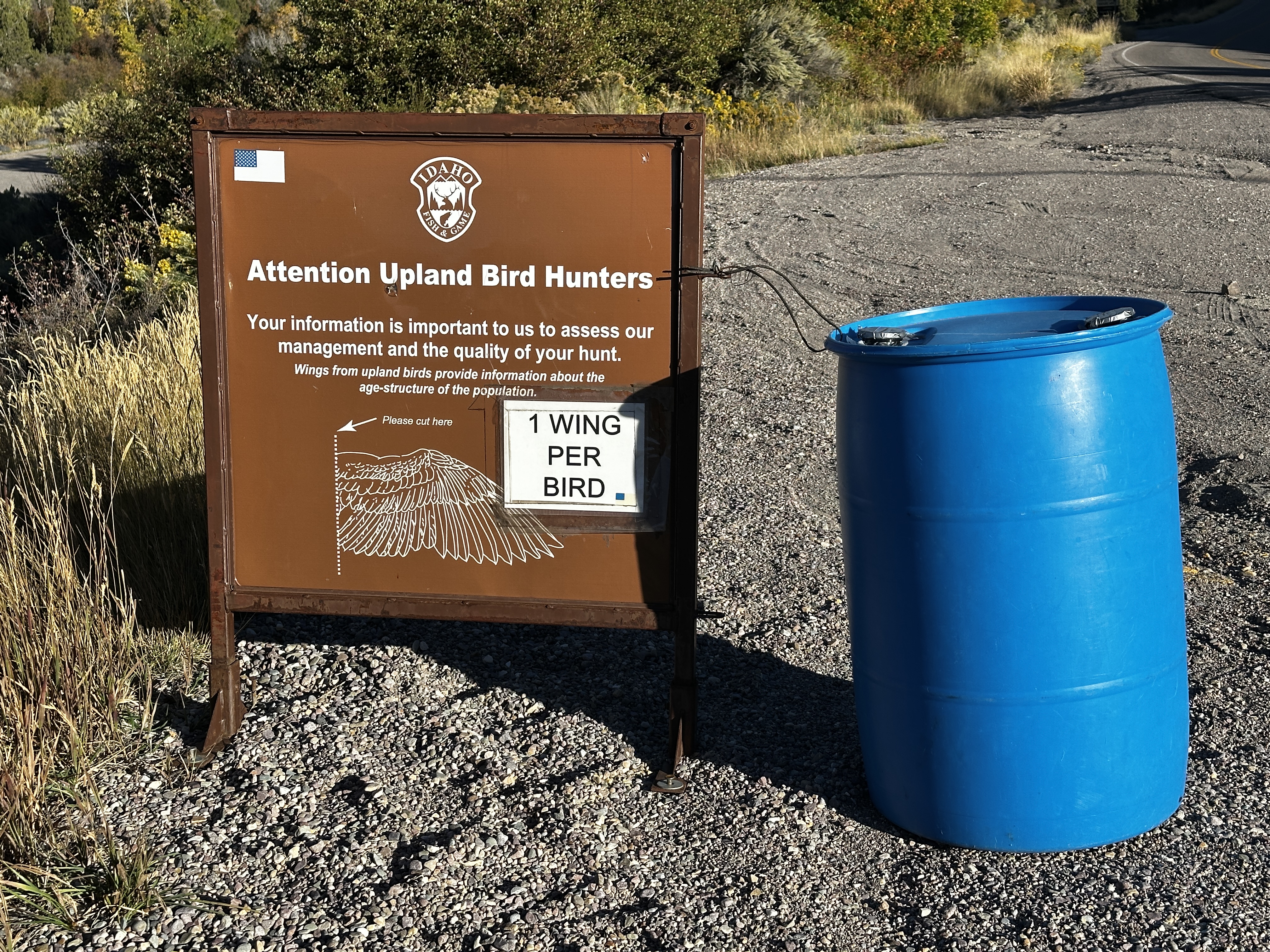 A blue metal barrel sits on a gravel parking area next to a large brown information sign