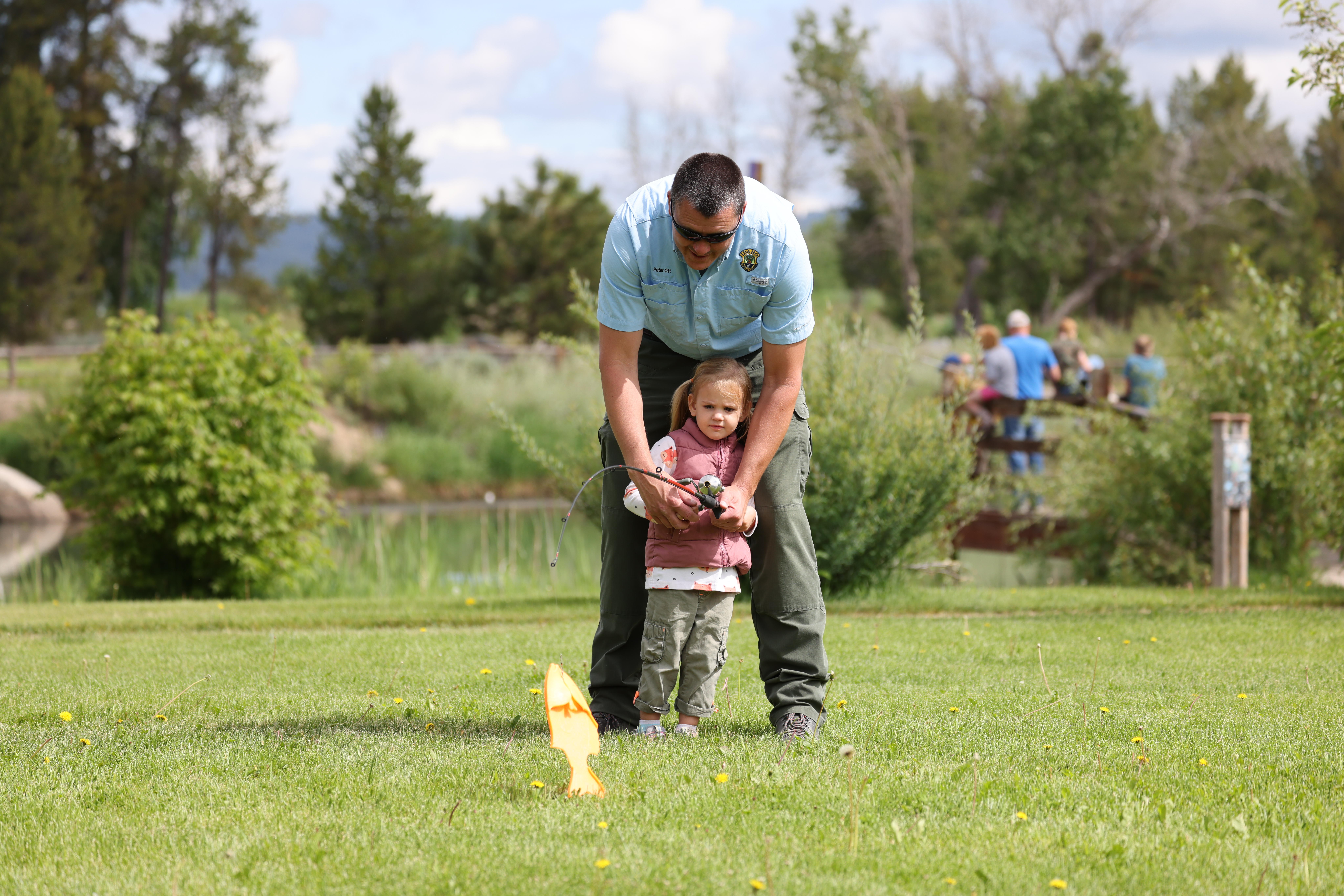 A Fish and Game employee helps a young girl reel in a plastic fish with a casting bobber.