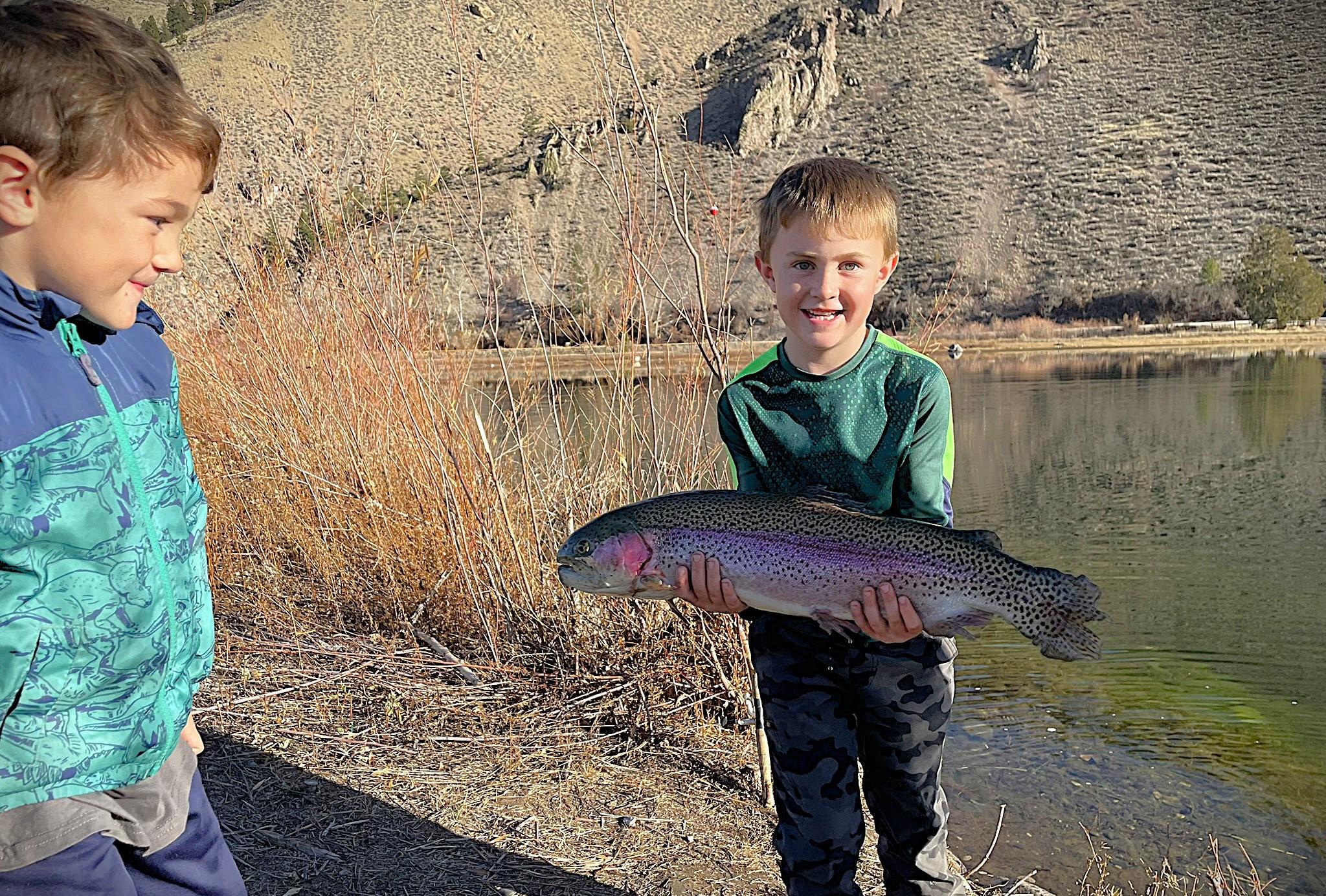 Young Boy holding a large rainbow trout near a pond as another young boy admires his catch.
