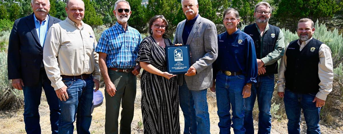 Group of people standing side by side outside with green trees in the background.  Woman in center is hold a plaque.