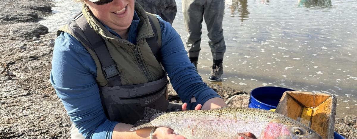 A woman in waders and cap holding a large rainbow trout with water and boat in the background