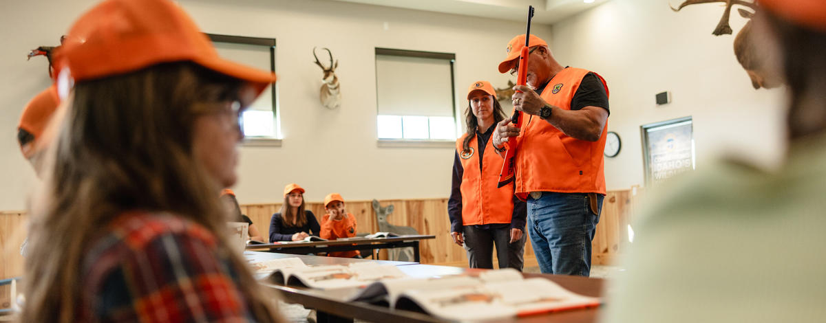 A hunter education instructor holding a firearm points at the trigger in front of a class.
