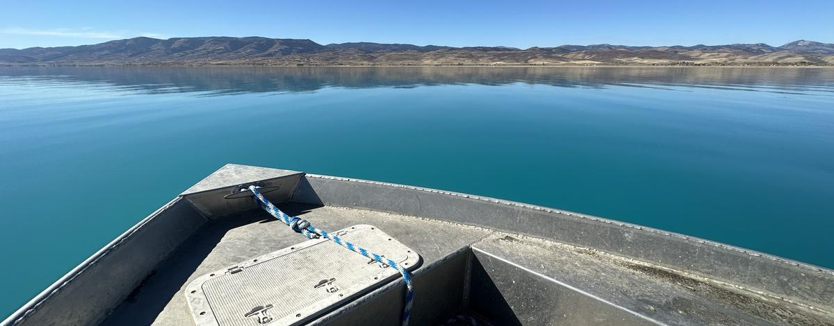 The front of a boat shown atop the bright blue waters of Bear Lake.  Foothills are in the background under a blue sky.