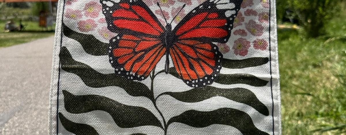 Flag featuring a black and orange monarch butterfly and the words "milkweed for monarchs" staked into a grassy area next to a path