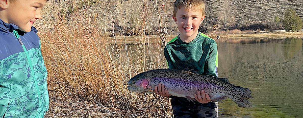 Young Boy holding a large rainbow trout near a pond as another young boy admires his catch.