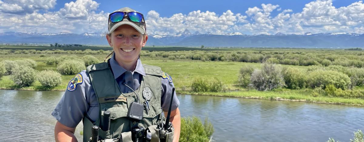 A conservation officer standing near a river and a scenic view behind her, smiles for the camera.