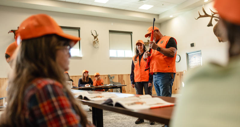 A hunter education instructor holding a firearm points at the trigger in front of a class.