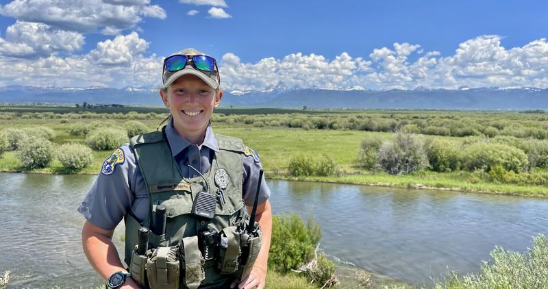 A conservation officer standing near a river and a scenic view behind her, smiles for the camera.