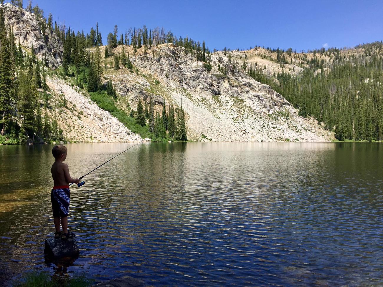 A boy wearing a swim suit fishes at an alpine lake.