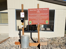 A garbage can next to a large sign with instructions asking mule deer hunters to deposit Chronic Wasting Disease samples in the can.