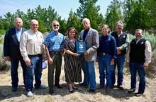 Group of people standing side by side outside with green trees in the background.  Woman in center is hold a plaque.