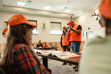 A hunter education instructor holding a firearm points at the trigger in front of a class.