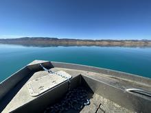 The bow of a jet boat projecting out onto a serene turquoise blue lake with distant mountains shown in the background.  A cloudless blue sky is shown above.