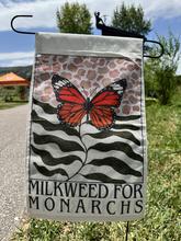 Flag featuring a black and orange monarch butterfly and the words "milkweed for monarchs" staked into a grassy area next to a path