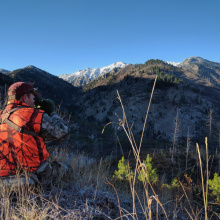 big game hunter dressed in hunter orange looking through binoculars on a mountain ridge Ben Studer medium shot, November 2016